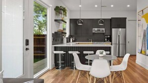 A modern kitchen with dark cabinetry, stainless steel appliances, and a central island. There are white bar stools and a round table with chairs. - Holly #B xxxxxxxxxxx (Austin)