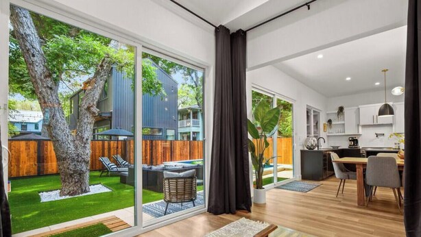 Un apartamento moderno con un amplio ventanal corredera que da a un patio trasero con un árbol y una terraza. En el interior, hay una cocina con armarios blancos y una zona de comedor con una mesa y sillas.