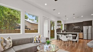A modern living room with a grey sofa, a glass coffee table, and a dining area with white chairs. There is a kitchen with dark cabinets and stainless steel appliances. - Brand New Luxurious Property + Huge Outdoor Space! (Austin)