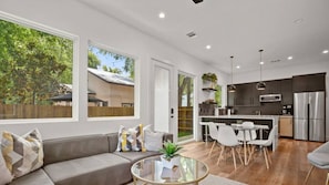 A modern living room with a grey sofa, a glass coffee table, and a dining area with white chairs. There is a kitchen with dark cabinets and stainless steel appliances.