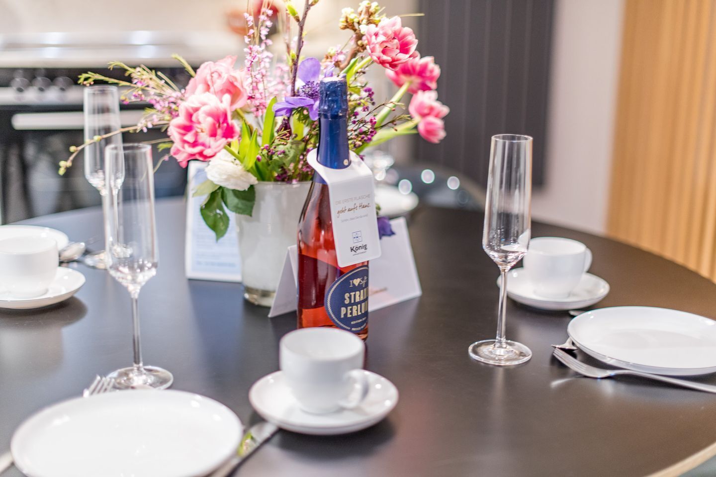 A dining table set with white plates, cups, and glasses, a bottle of wine, and a floral centerpiece.