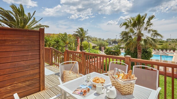 Une terrasse en bois avec des sièges d’extérieur, une table dressée avec des aliments pour le petit-déjeuner, et une vue sur une piscine et des palmiers.