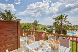 A wooden deck with outdoor seating, a table set with breakfast items, and a view of a swimming pool and palm trees. - Appartement Côté Piscine + Balcon/Terrasse (Agde)