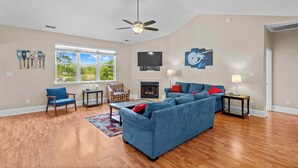 Ein geräumiges Wohnzimmer mit Holzdielen, einem blauen Sofa, einem gemusterten Sessel, einem Kamin und einem Deckenventilator.