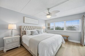 Ein Schlafzimmer mit einer geblatteten Kopfteil, einem Bett mit weißer Bettwäsche, einem Nachttisch mit einer Lampe, einem Deckenventilator und einem Fenster mit Jalousien.