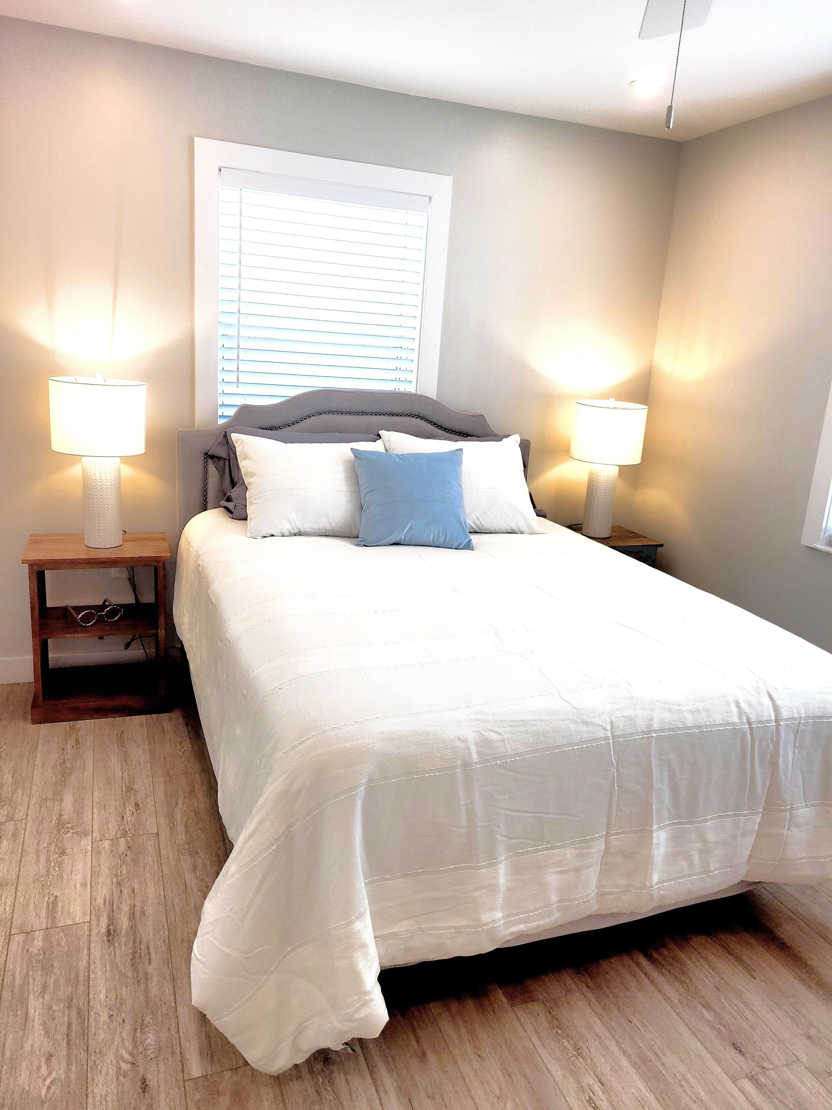 A neatly made bed with white bedding and two pillows, flanked by two bedside tables with lamps, in a room with wooden flooring and a window with blinds.