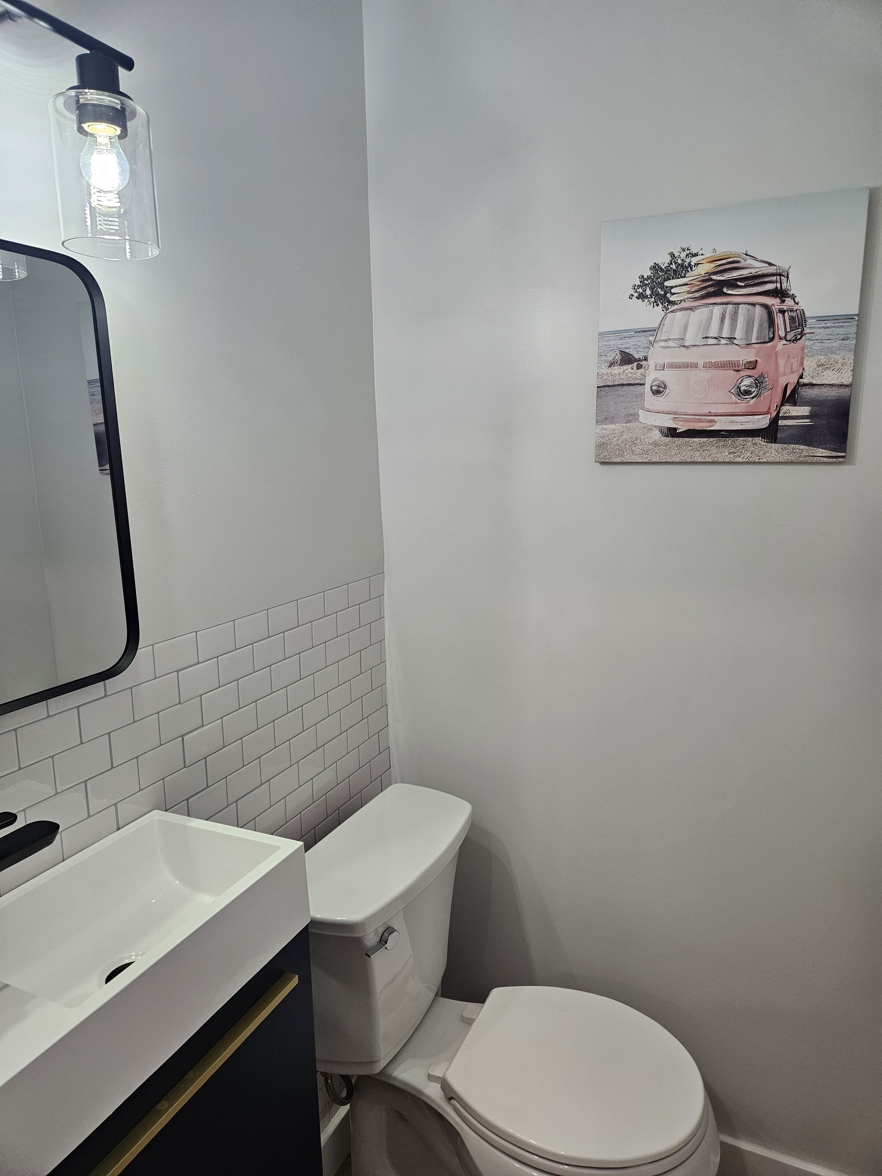 Un bagno con un WC bianco, un lavandino bianco e uno specchio. Sul muro, un quadro raffigura un furgone rosa.