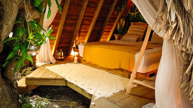 Une chambre à coucher chaleureuse et rustique, avec un plafond en bois, un lit recouvert d’une courtepointe jaune et une petite plateforme en bois sur laquelle sont posées des bougies.