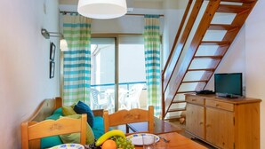 A dining area with a wooden table set for two, a fruit bowl, and a television on a wooden cabinet.