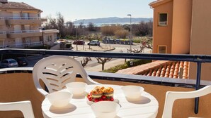 A balcony with a round table and chairs, a dessert on the table, and a view of a parking lot and buildings in the background.