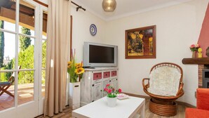 A living room with a television, a chair, a table with flowers, and a view of the outdoors. - Stunning  villa for 4 people with pool and terrace (Torroella De Montgrí)
