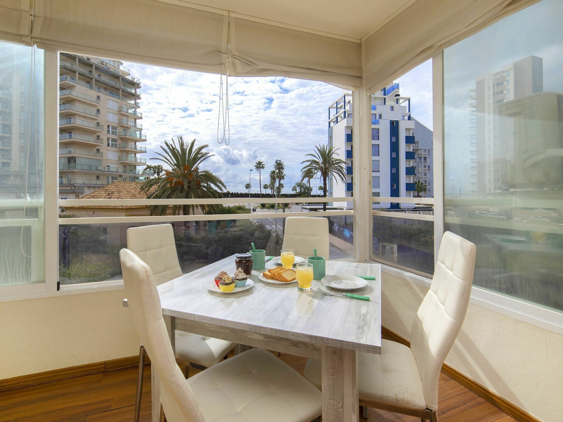 Un coin repas avec une table dressée pour deux personnes, une vue sur le paysage urbain et un balcon agrémenté de palmiers.