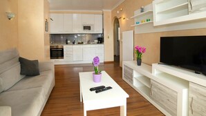 Un salon moderne avec un canapé, une table basse ornée d’un vase de fleurs, un téléviseur à écran plat et une cuisine en arrière-plan.