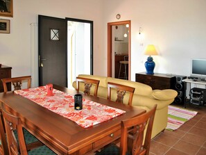 Un'area pranzo con tavolo in legno, sedie e un divano. Si trova una televisione, degli altoparlanti e una lampada su un tavolo.