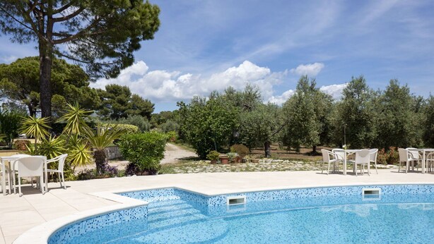 A swimming pool with a tiled edge, surrounded by white chairs and tables, and a garden with trees and plants. - Cosy apartment for 6 people with WIFI, pool, TV, patio and pets allowed (Sperlonga)