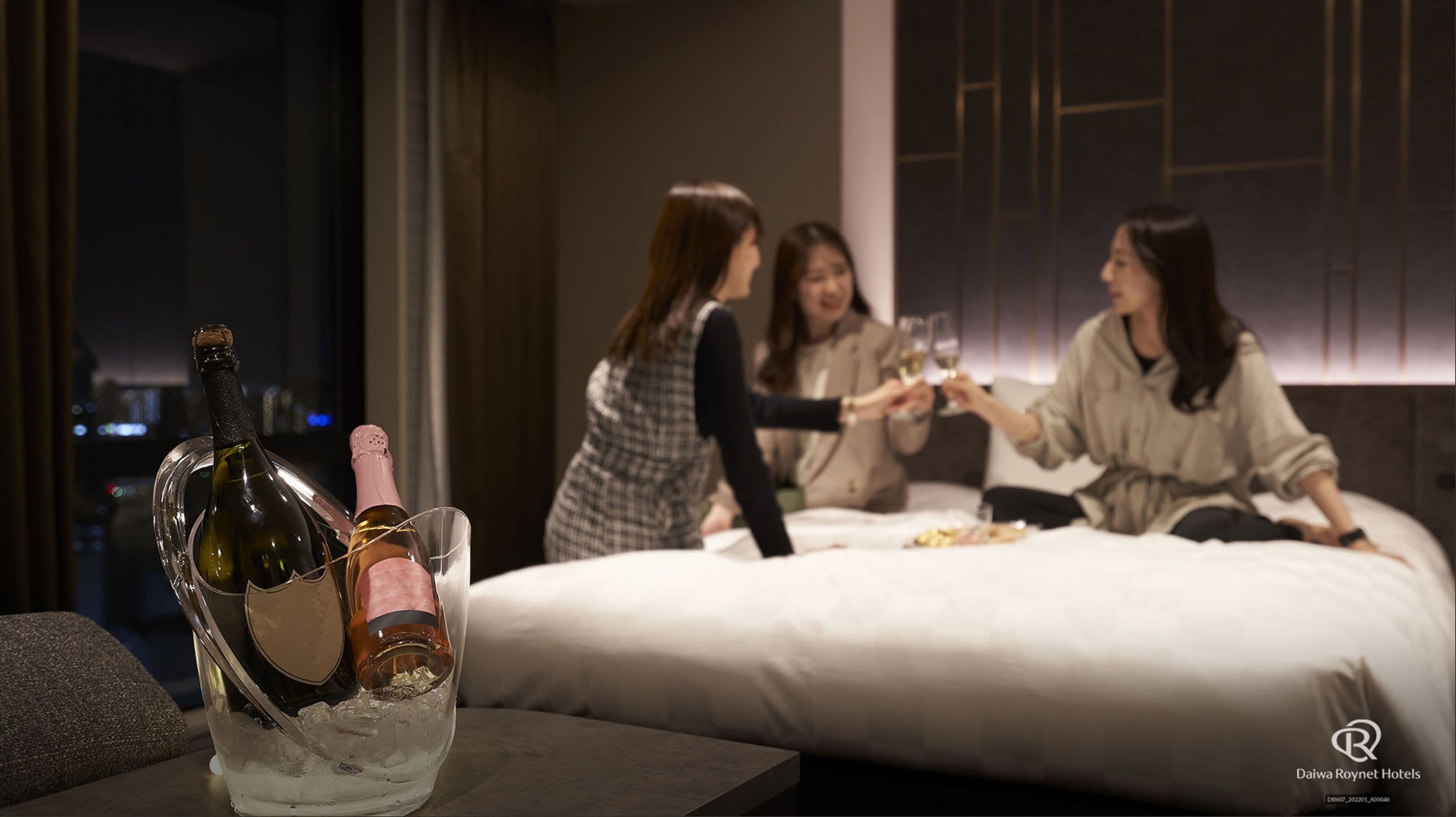A hotel room with a bed, a table with a bottle of champagne, and a city view.