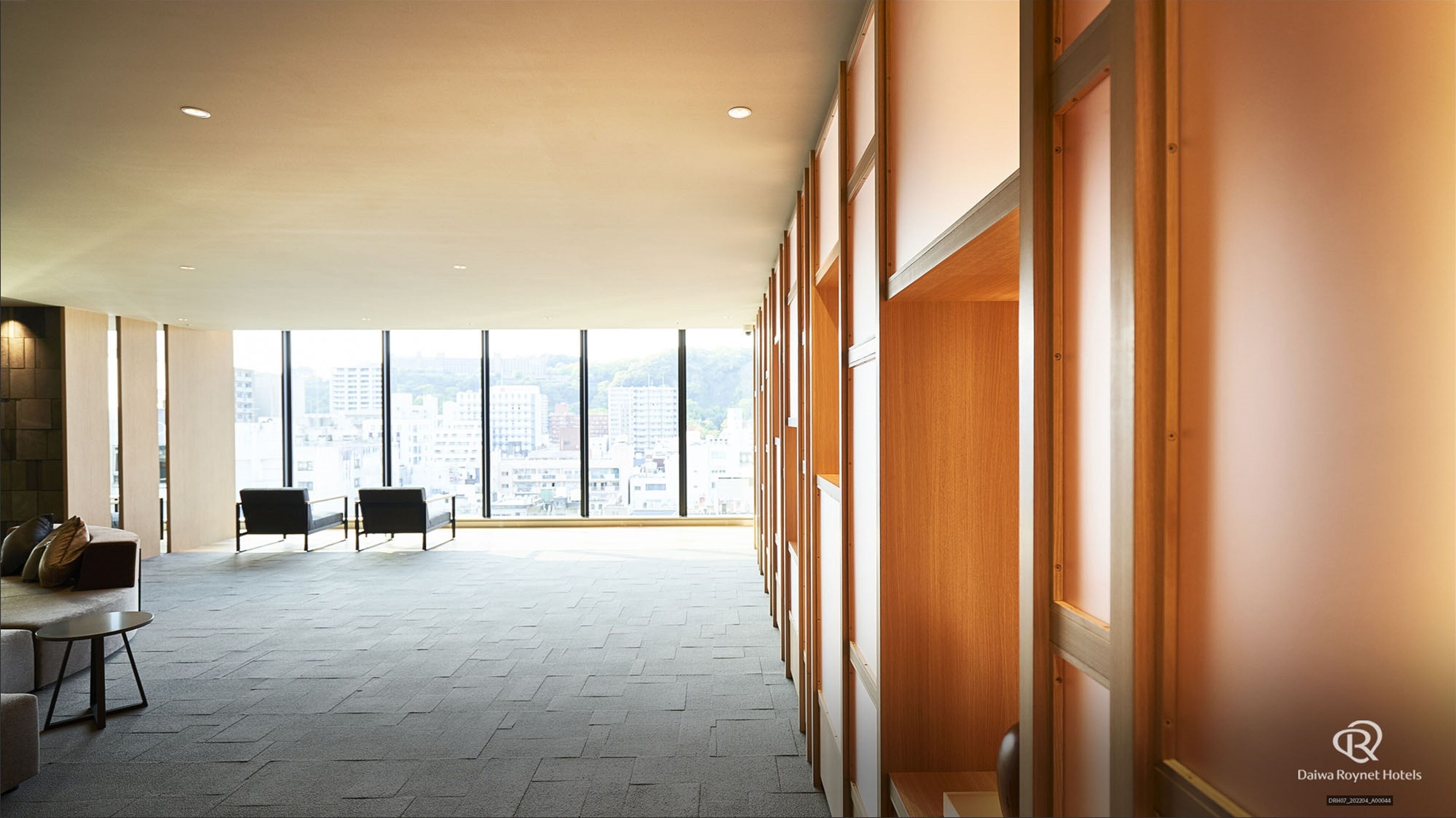 A spacious hotel lobby with high ceilings, large windows offering city views, and a seating area with sofas and a small table.