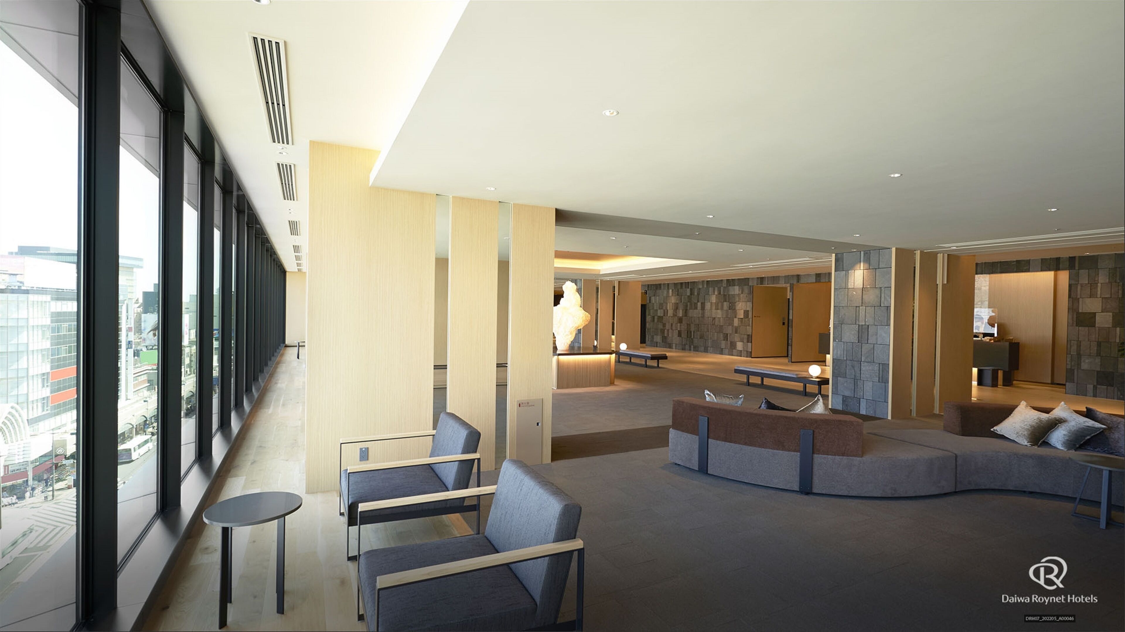 A modern hotel lobby with seating areas, a reception desk, and a view of the cityscape.