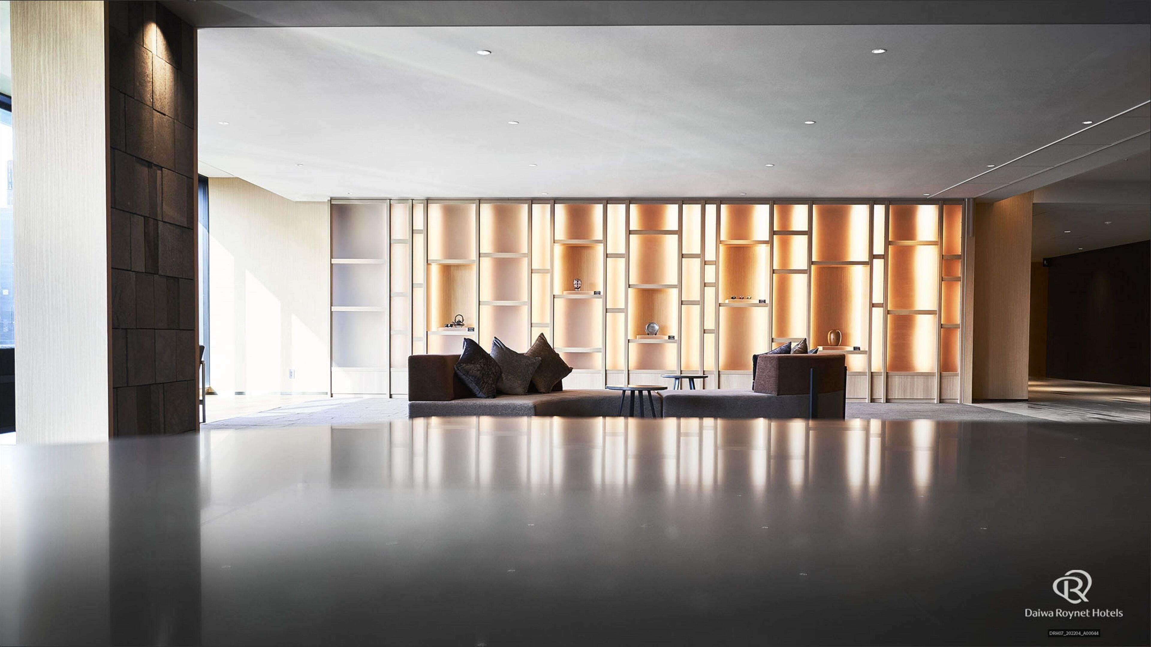 A spacious lobby with a modern design, featuring a seating area with a sofa and armchairs, a coffee table, and a small table with a lamp. The walls have built-in shelves with decorative items.