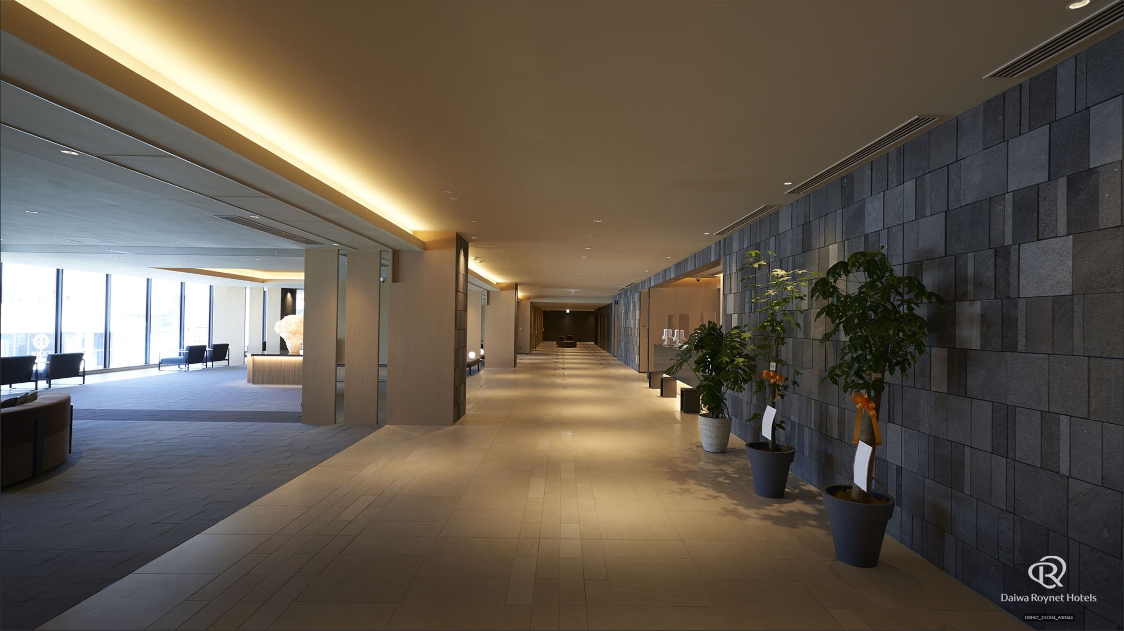 A modern hotel lobby with a long corridor, seating areas, and potted plants.