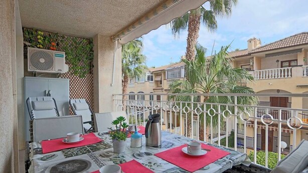 Een balkon met een tafel voor twee personen, een plafondventilator en uitzicht op omliggende gebouwen en palmbomen.