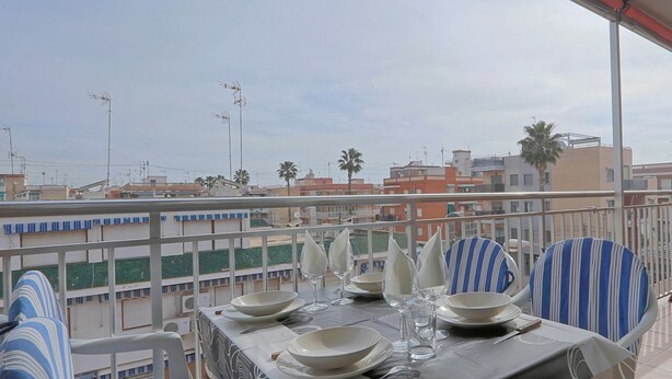 A balcony with a table set for dining, overlooking a residential area with buildings and palm trees. - 060 - Gran Playa 004 - comfortHOLIDAYS (Santa Pola)