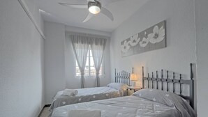 A bedroom with two beds, a ceiling fan, a window with curtains, and a wall-mounted artwork. - 060 - Gran Playa 004 - comfortHOLIDAYS (Santa Pola)
