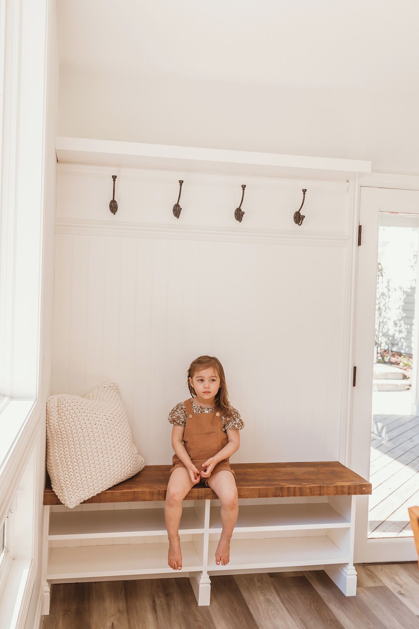 A child sitting on a wooden bench in a room with a white wall and a white door.