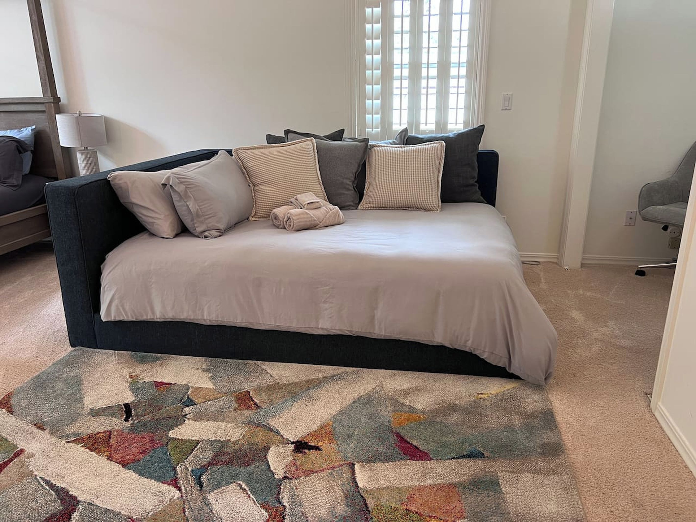 A bedroom with a dark-colored sofa bed, a patterned rug, and a window with blinds.