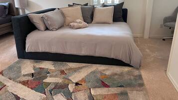 A bedroom with a dark-colored sofa bed, a patterned rug, and a window with blinds.