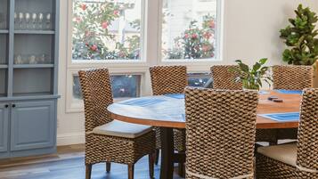 A dining area with a round table and wicker chairs, a blue and white patterned tablecloth, a wooden ceiling, and a large window with a view of trees and buildings.