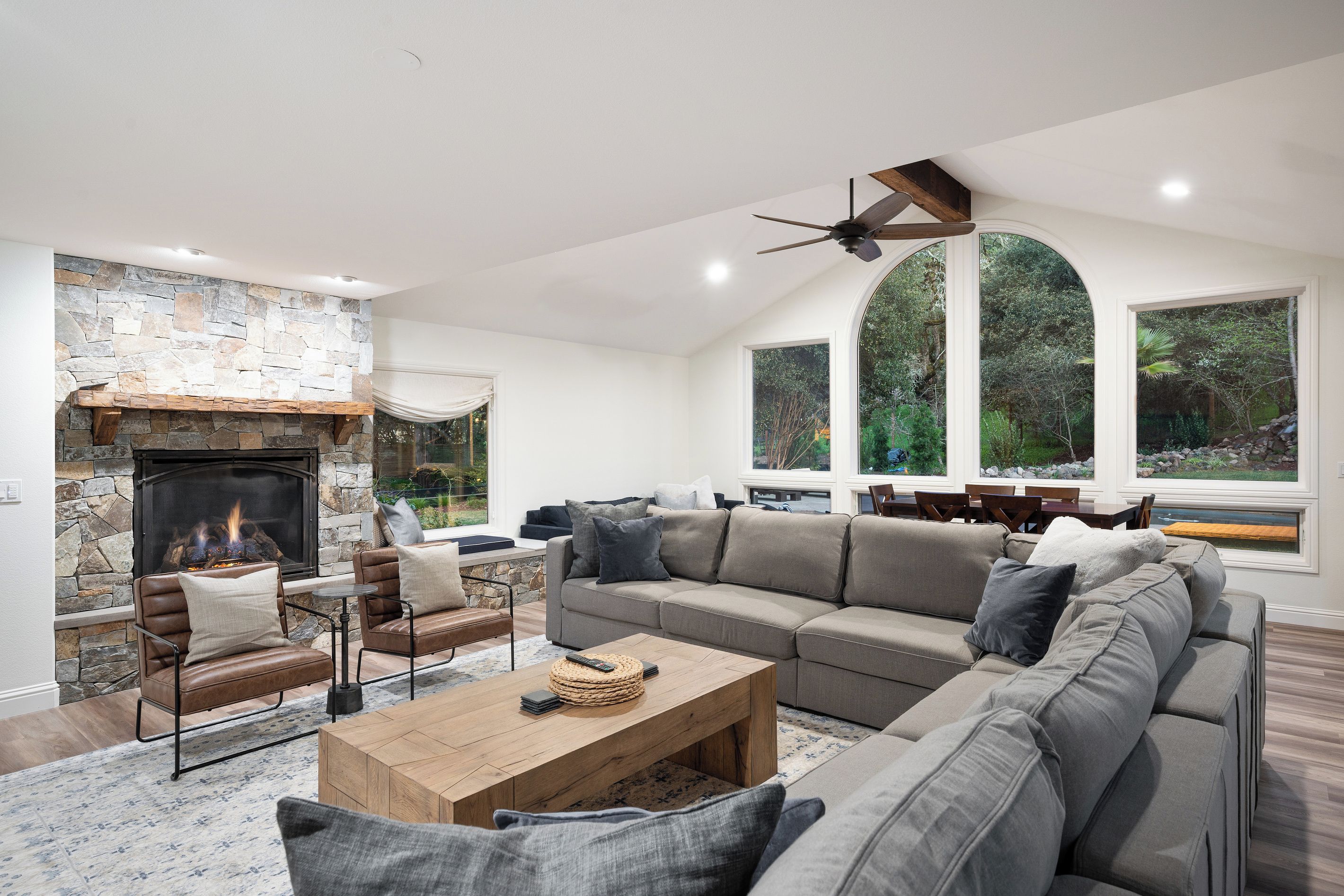 A modern living room with a stone fireplace, leather sofas, a wooden coffee table, and large windows offering a view of the outdoors.