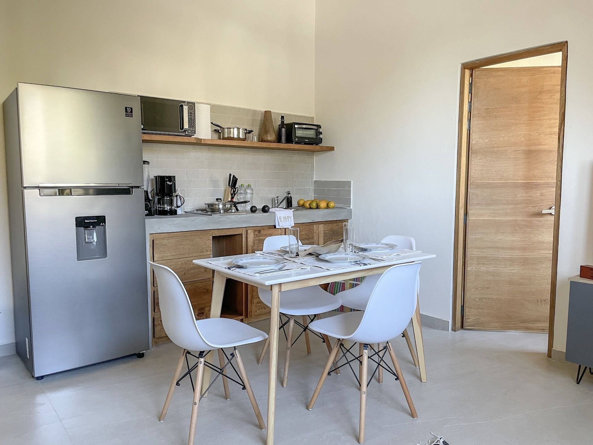 Uma cozinha compacta com geladeira, micro-ondas e cafeteira. Há uma mesa de jantar com cadeiras e uma porta de madeira ao fundo.