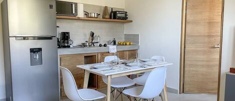 Cocina compacta con refrigerador, microondas y cafetera. Hay una mesa de comedor con sillas y una puerta de madera al fondo.