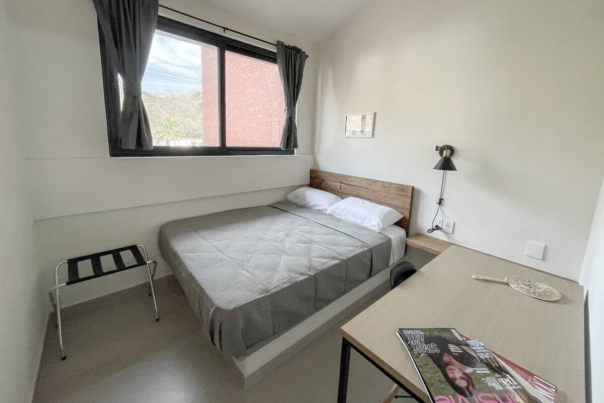 Une chambre avec un lit simple, une fenêtre, une table de chevet, une lampe et un magazine posé sur la table.