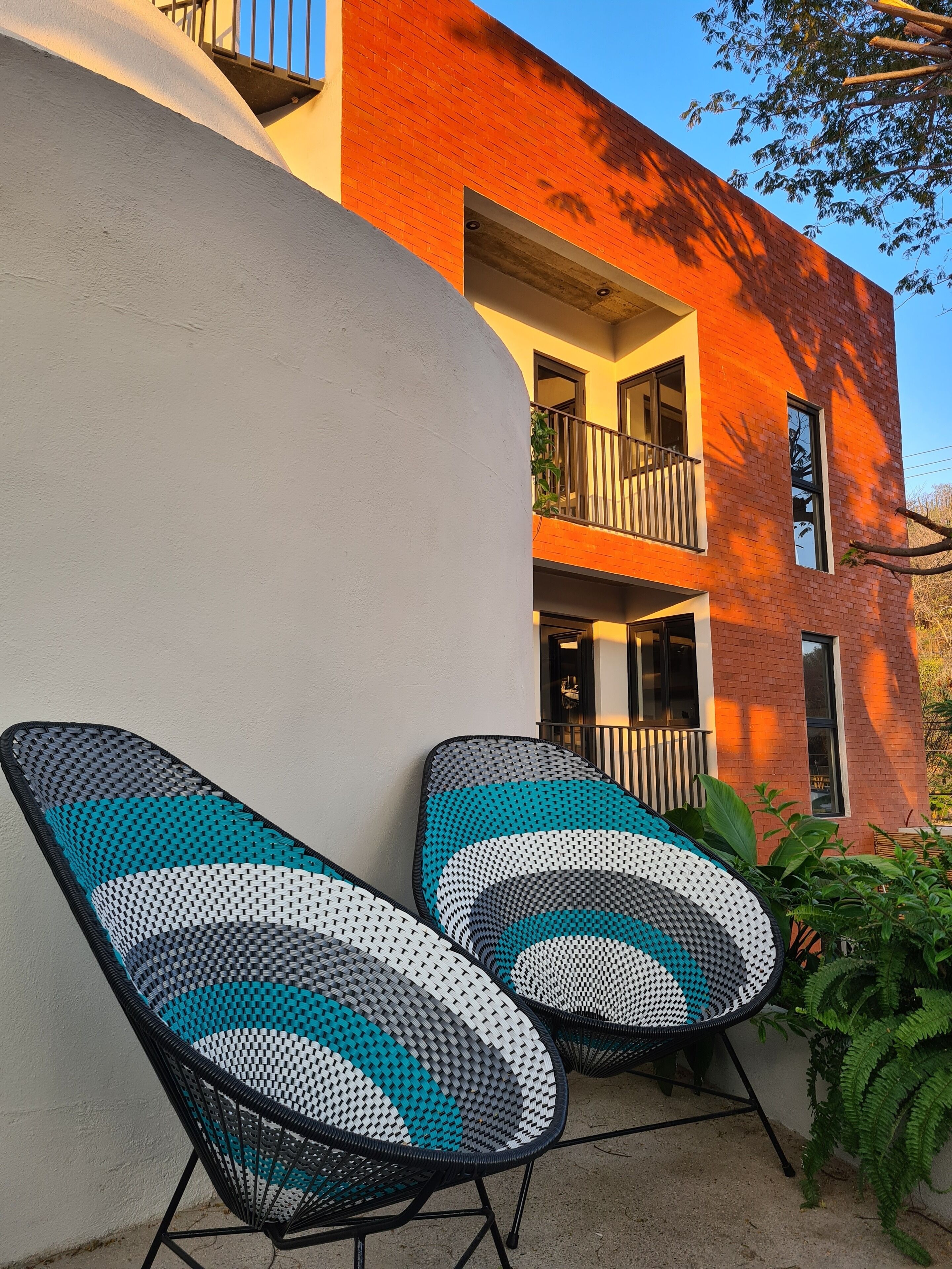 Deux fauteuils de jardin à motifs à carreaux, placés sur un balcon d’où l’on peut admirer un immeuble en briques rouges et un ciel d’un bleu éclatant.