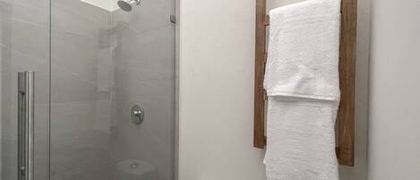 A bathroom with a glass-enclosed shower, a toilet, and a wooden towel rack with two white towels.