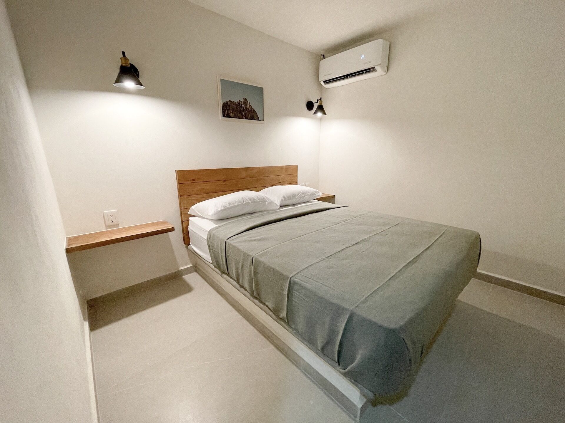 Un dormitorio ordenado con cabecera de madera, cama individual con colcha gris, mesita de noche, aire acondicionado de pared y un pequeño marco de fotos.