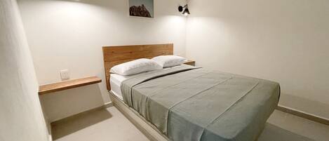 Un dormitorio ordenado con cabecera de madera, cama individual con colcha gris, mesita de noche, aire acondicionado de pared y un pequeño marco de fotos.