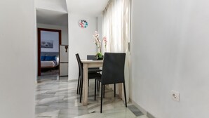 A dining area with a table, chairs, and a vase of flowers. A bed and a refrigerator are visible in the background.