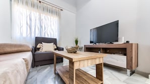 A modern living room with a sofa, a wooden coffee table, a TV on a wooden stand, and a large window with sheer curtains.