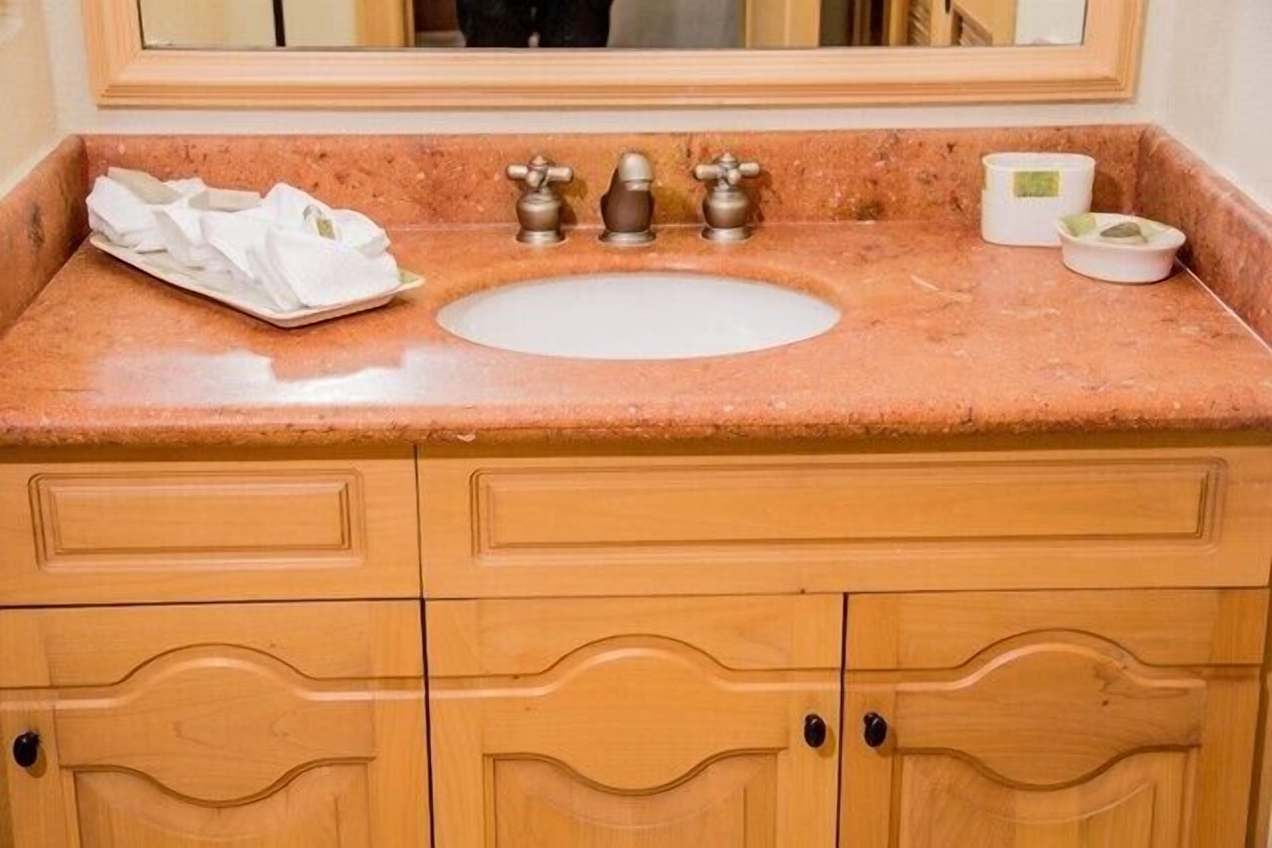 Une salle de bain avec un lavabo en marbre, trois robinets et un miroir au-dessus. On trouve deux petits récipients sur le comptoir du lavabo.