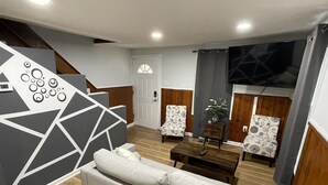 Una sala de estar moderna con un sofá gris, piso de madera, una escalera con arte mural geométrico y un televisor al fondo.