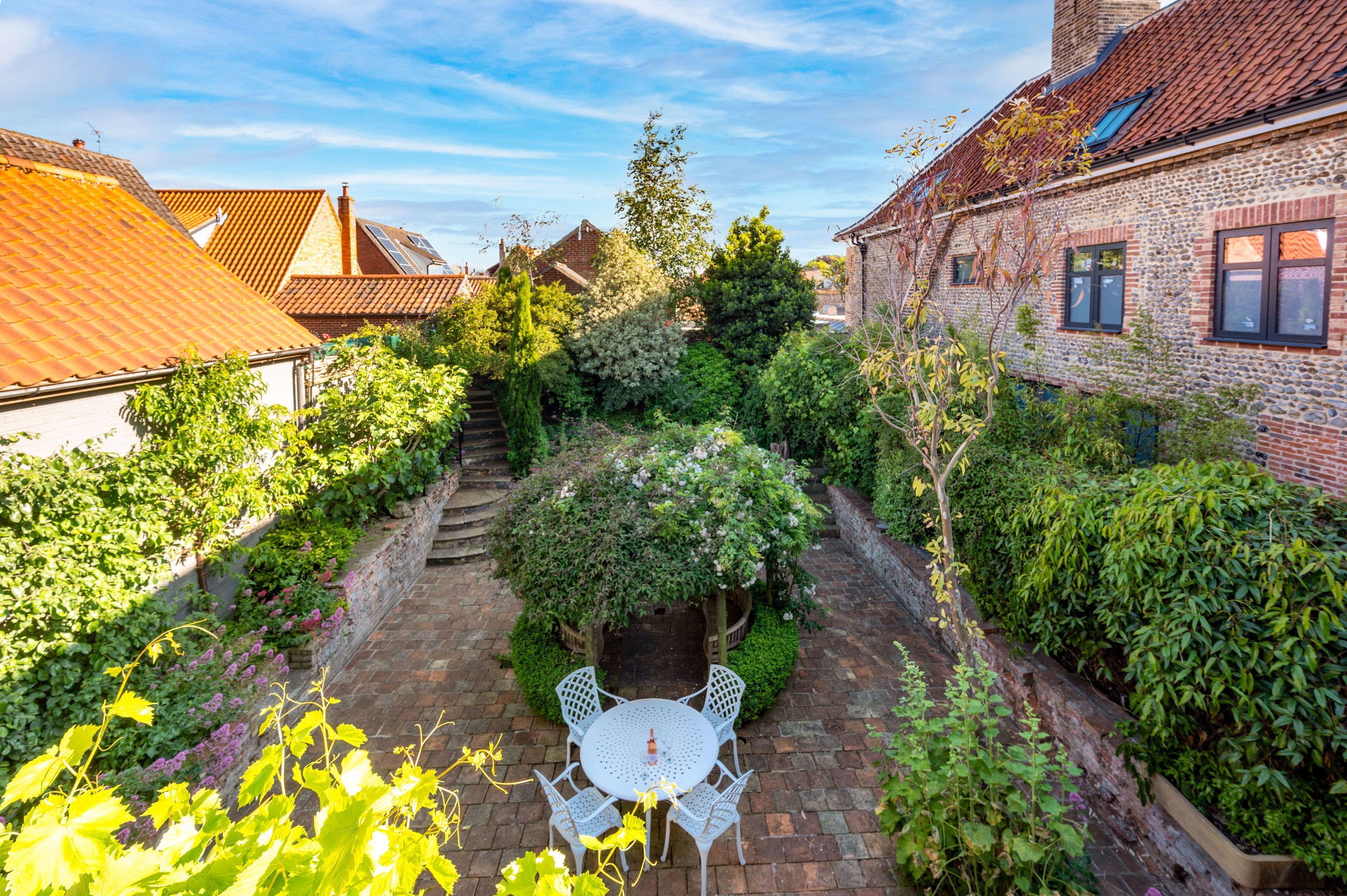 Une cour avec un chemin en briques, des meubles d’extérieur blancs et une végétation luxuriante.