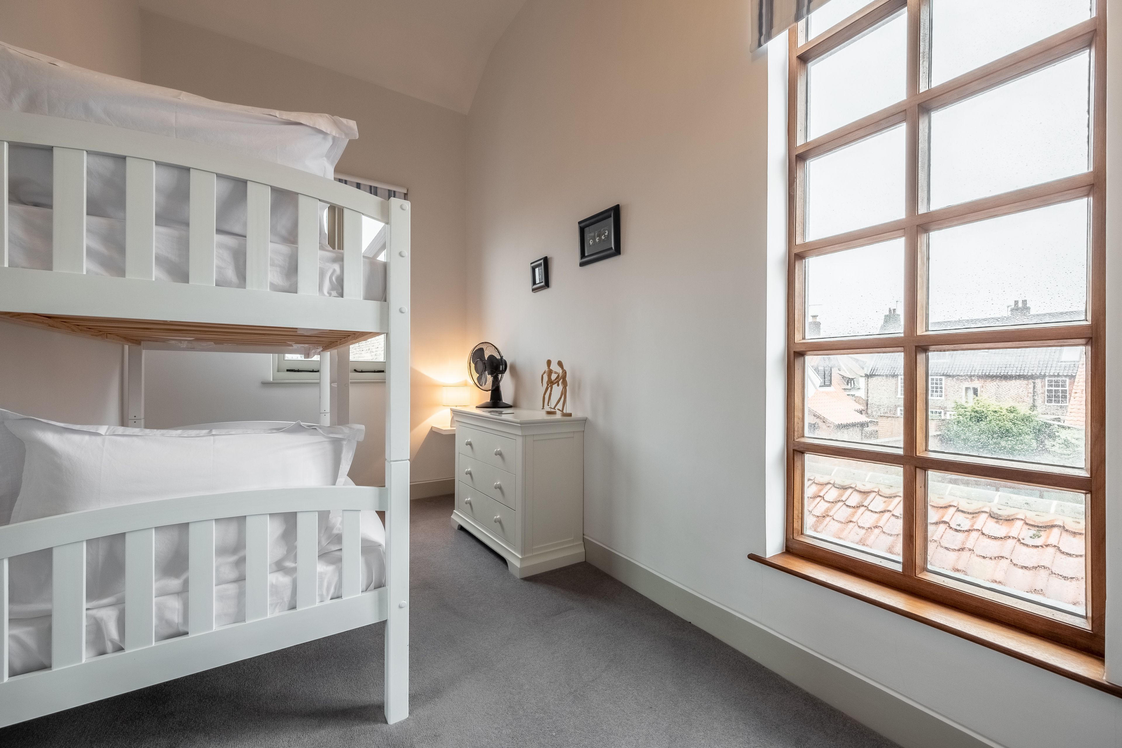 A room with bunk beds, a bedside table with a lamp, a dresser, and a window with a view of buildings.