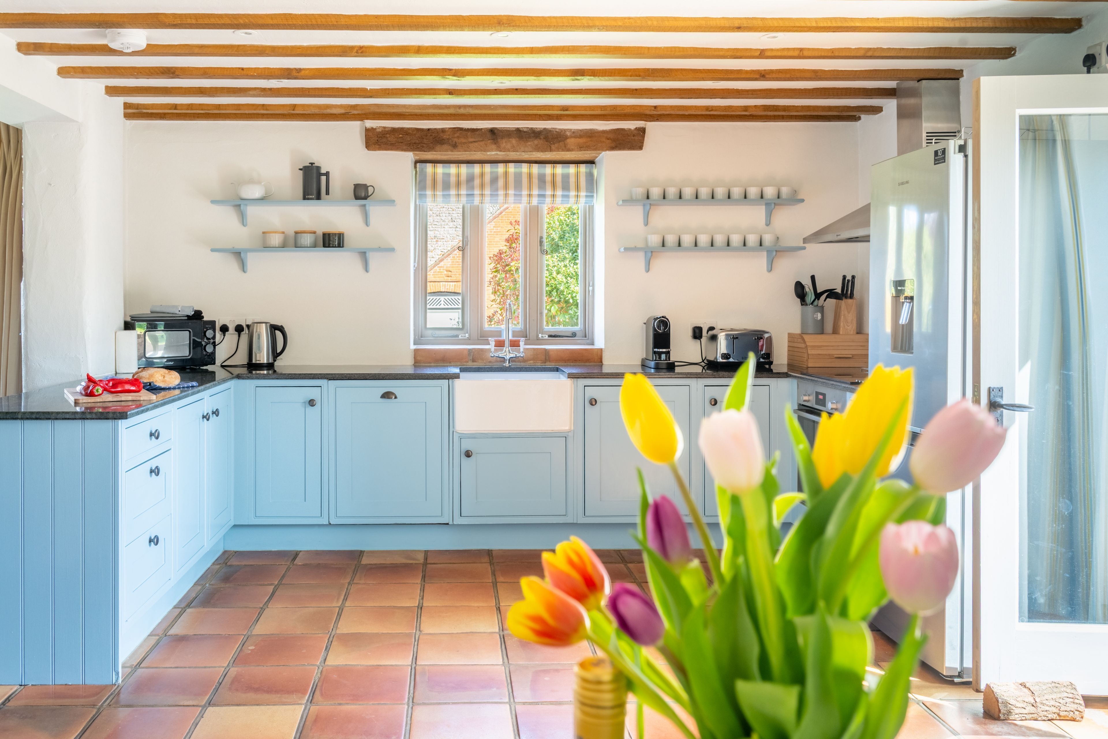 A kitchen with light blue cabinets, a tiled floor, a window with a view, and a refrigerator.