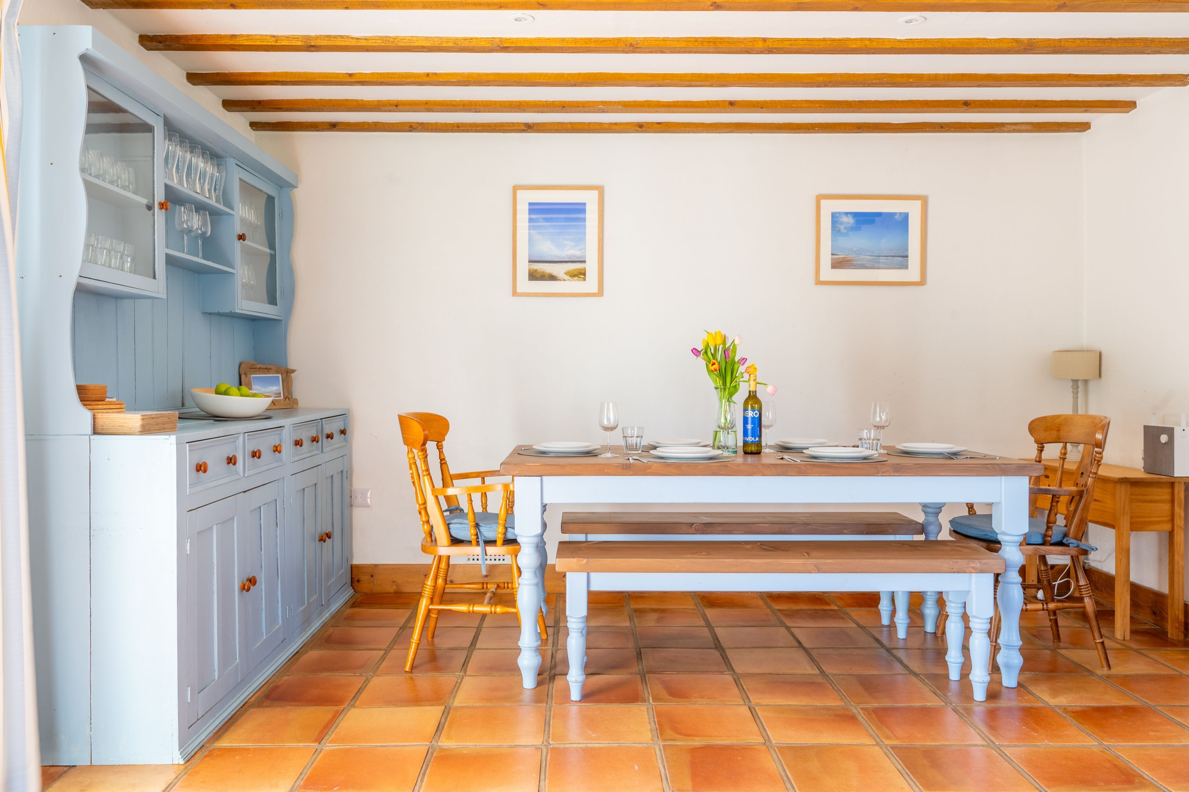 A dining area with a wooden table set for four, surrounded by chairs. A blue cabinet with glass doors is to the left, and a wooden bench is in front of the table.