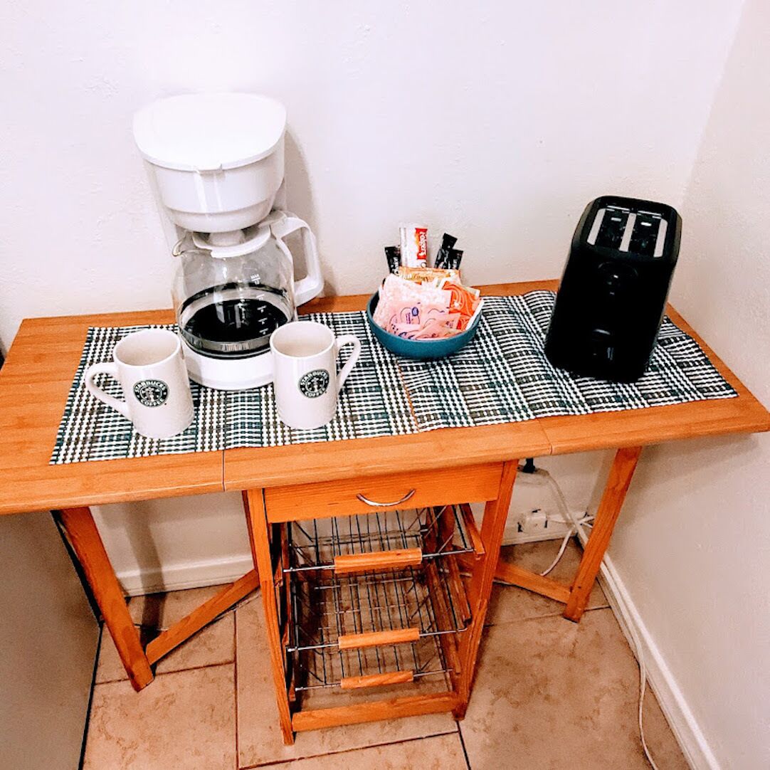 A small wooden table with a checkered placemat, two white coffee mugs, a coffee maker, a blue bowl with snacks, and a black speaker.