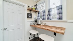 Una cucina con una porta bianca, una finestra con tende, uno scaffale in legno pieno di oggetti da cucina e due sgabelli da bar bianchi.