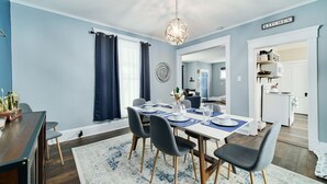 Una sala da pranzo con un tavolo apparecchiato per quattro persone, un lampadario e una zona cucina visibile attraverso un'apertura.